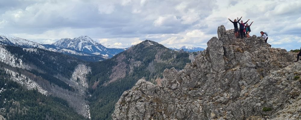 Kids trekking at the top of a rocky mountain with snowy tree covered mountains in the distance.