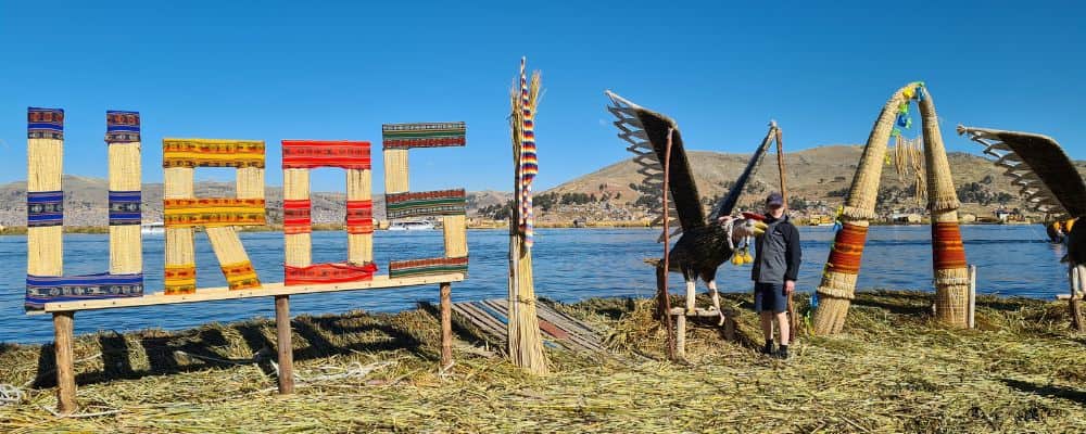 A kid stood next to a large bird statues and 'UROS' word near a lake in the sunshine. Another perfect place to stop on a family gap year. 