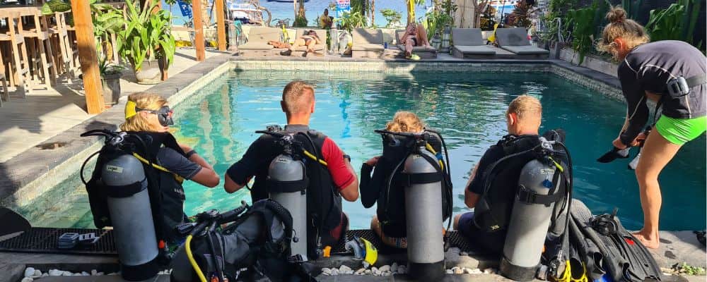 A family sat at a pool in a tropical destination with scuba diving kit on in the sun. A great gap ear activity.