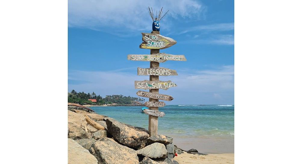 A surf school sign post on the beach by the sea offering surf camps Sri Lanka.