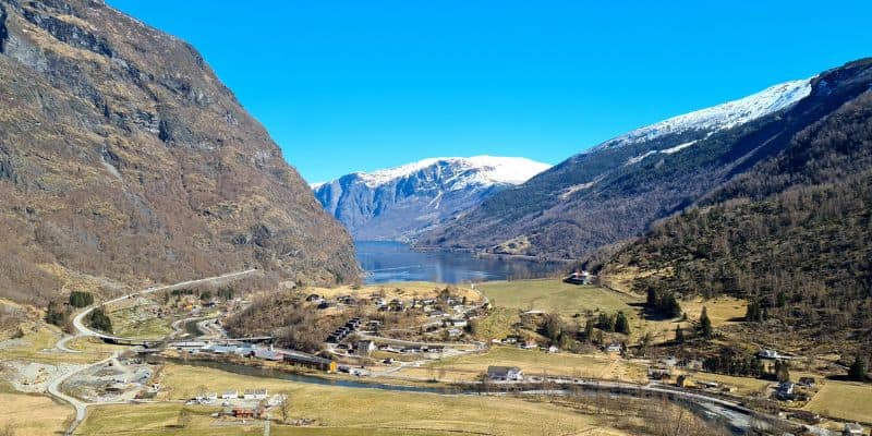 View of green fjords on a sunny day with blue skies and snow capped peaks in the distance. A village and road with cars.