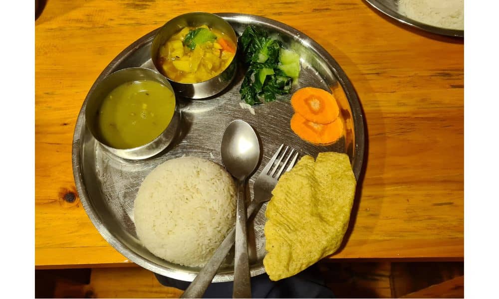 A metal plate on a wooden table with knife, folk, rice, soup, curry, carrot and a poppadom. A traditional meal on Everest Base Camp.