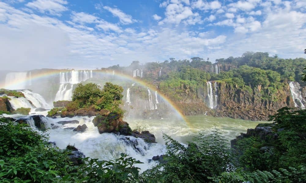 Multiple cascading waterfalls over rocks surrounded by forest and the best view of a rainbow.
