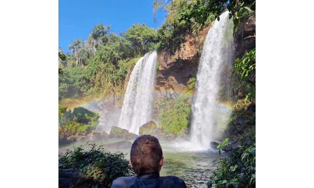 a kid viewing two waterfalls tumbling over a cliff surrounded by rainforest with a rainbow and blue sky when visiting Iguazu.
