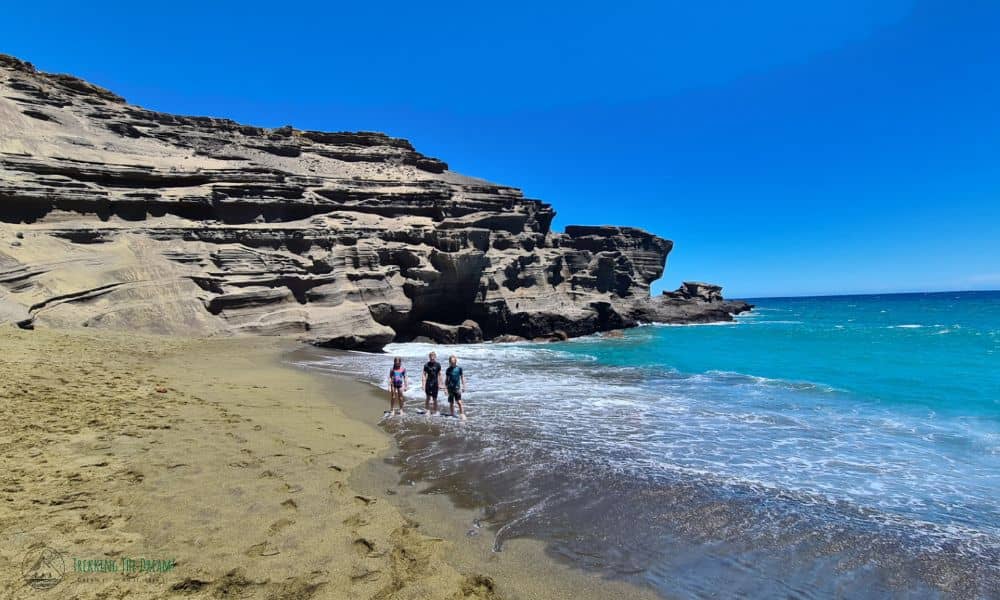 kids stood in the ocean at Papakolea beach with green sand, volcanic rock and blue sky.