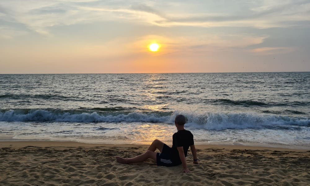 A kid sat on a yellow beach in Sri Lanka looking out to rolling waves in the ocean where the yellow sun is setting.