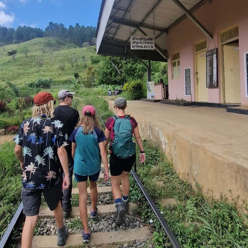 A family of four hiking on the rail track past Kithalella Railway Station on the way to Ella Rock view point surrounded by green hills and blue sky.  