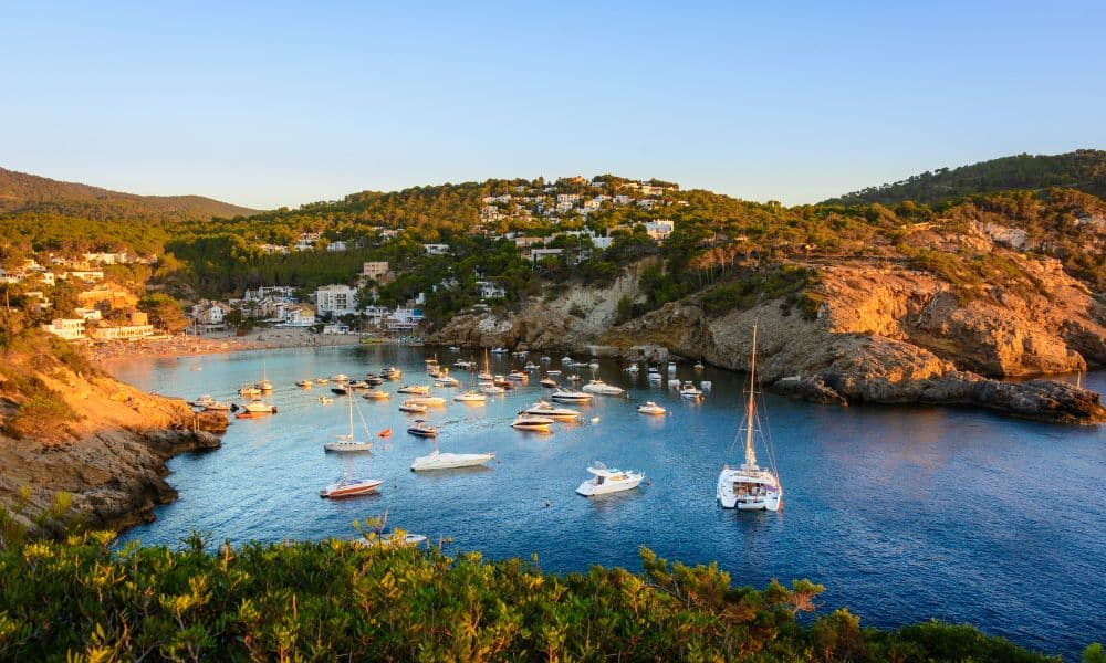 Near and far cliffs covered in green moss with blue ocean in a bay and lots of white sail boats floating in Cala Vedelle, Ibiza, Spain.