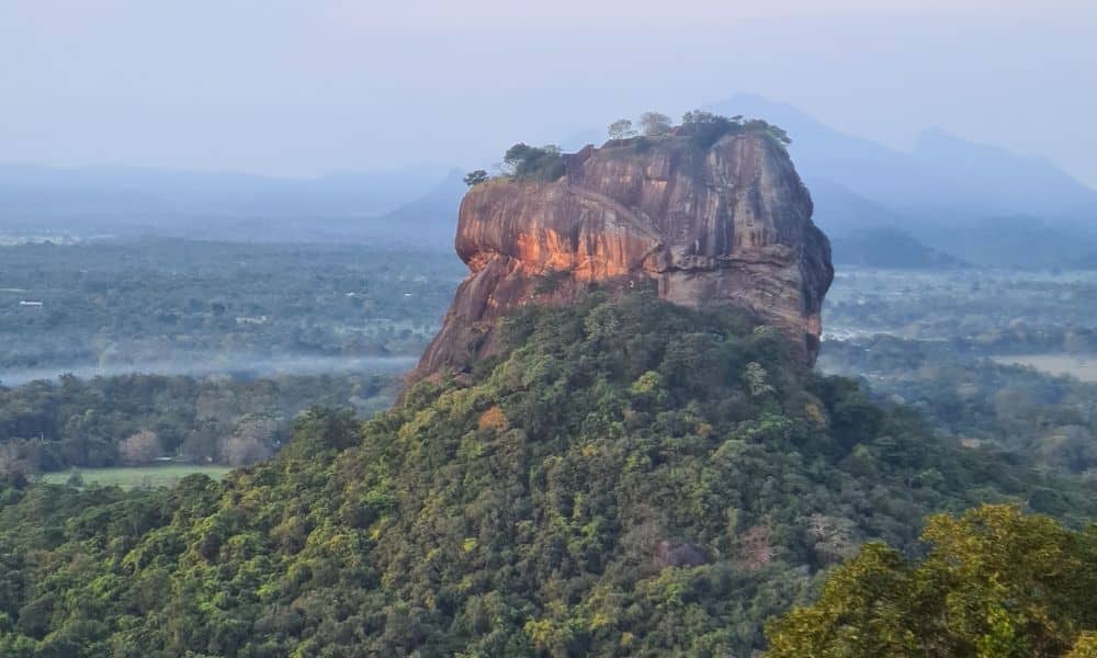 A large rock formation sticking out of the forest, surrounded by trees and mountains in the background. Climbing Sigiriya Rock is one of the tope things to do. 