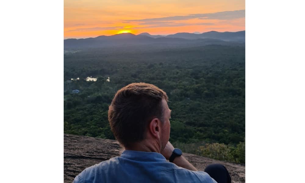 Back of a man sat on a rock overlooking green jungle as an orange sun rises behind hills creating an orange sky. This is the view from Pidurangala.