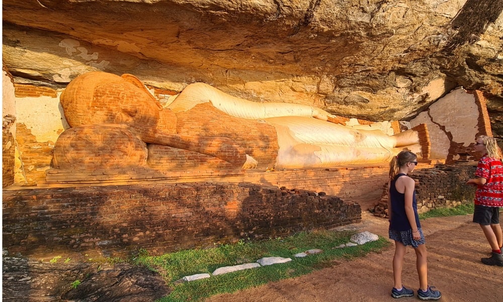 The sun is shining on a 12m reclining brick buddha within a rock face. Two kids are viewing during their Pidurangala hike.