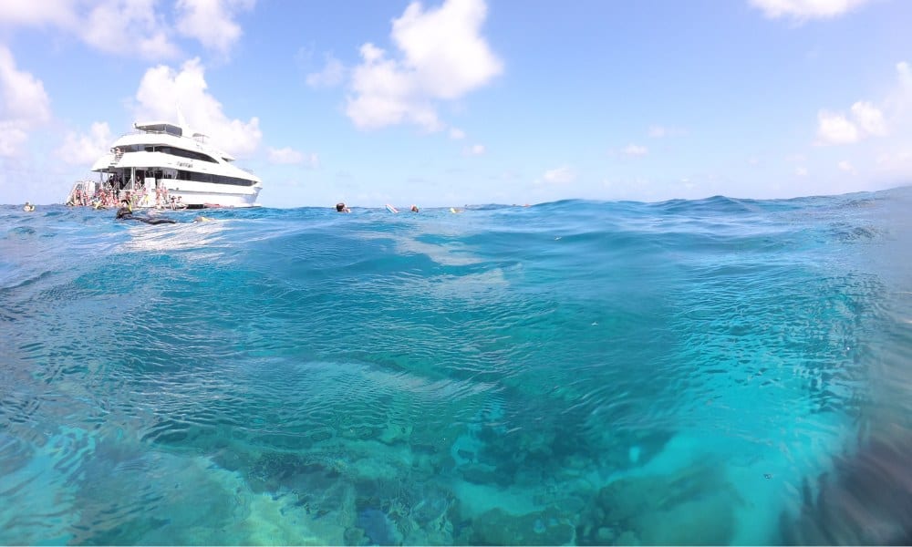 A white boat on the sea over an underwater reef with blue skies and fluffy white clouds. Snorkeling at the Barrier Reef is a top thing to do in Australia.