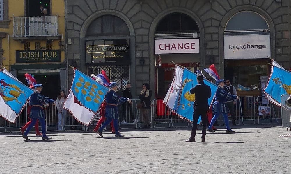 People parading with large blue and white flags in front of shops in Florence, Italy with families watching.