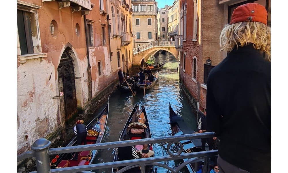 A narrow canal between buildings with bridges and gondolas carrying people. A kid is watching this popular activity in Venice, Italy 