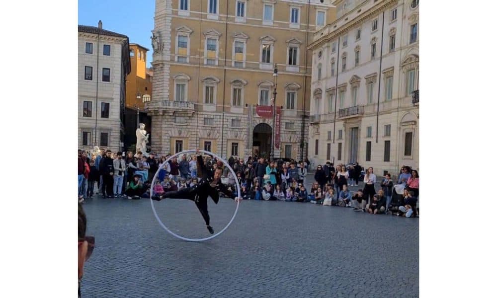 A crowd of people watching a man perform in a large hoop in an open court surrounded by tall buildings in Rome, Italy.