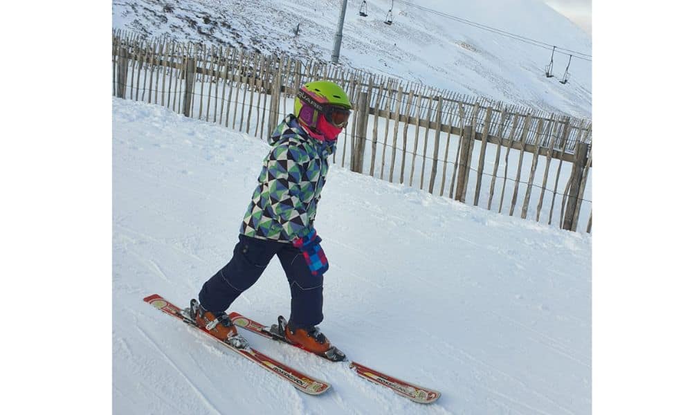 Trekking The Dream kid skiing downhill with gondolas in the distance. Cairngorm mountains, Scotland in winter