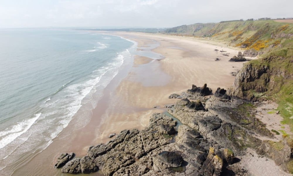 long, wide stretch of yellow sandy beach with a rocky formation. Blue sea rolling in. A classic coastal scene in Scotland