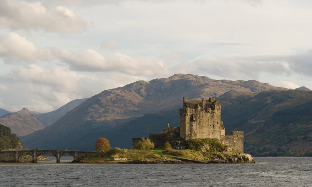 A stone castle on its own island with bridge access over water and scottish hills in the background. One of the famous castles in Scotland.