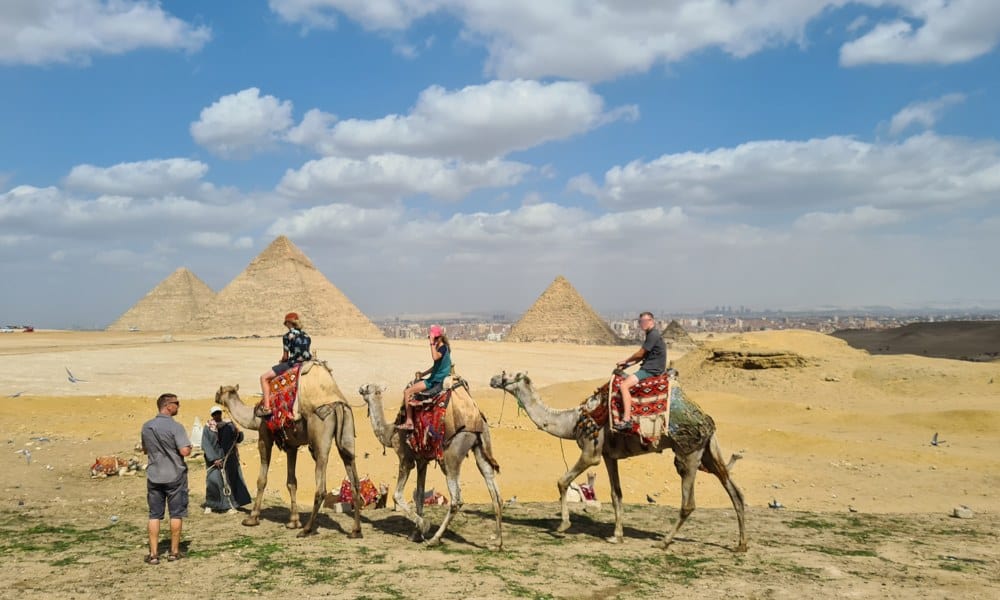 Trekking the Dream Family in the desert with three pyramids in the distance. Kids are riding camels. 