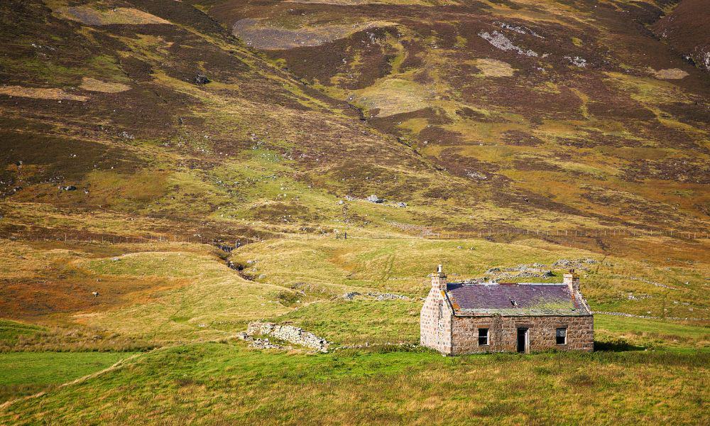 A remote derelict stone building surrounded by vast green fields and hills in Cairngorms, Scotland.