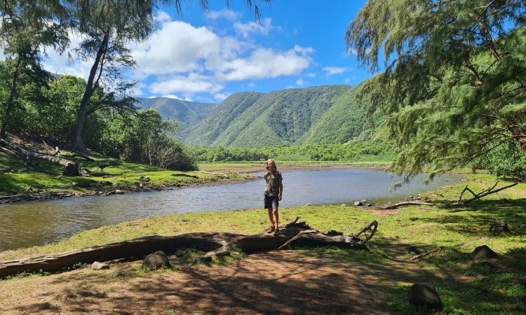 Kid stood in the shade by a river surrounded by a green valley. Pololū Valley is a popular hike as part of a Big Island itinerary.