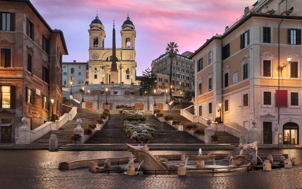 Break of dawn with purple sky behind Roman buildings, steps and a fountain. The Spanish Steps are a central attraction in Rome, Italy.