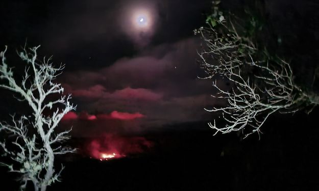 Red glow from a volcano in the distance with a moon in the night sky at a National Park.