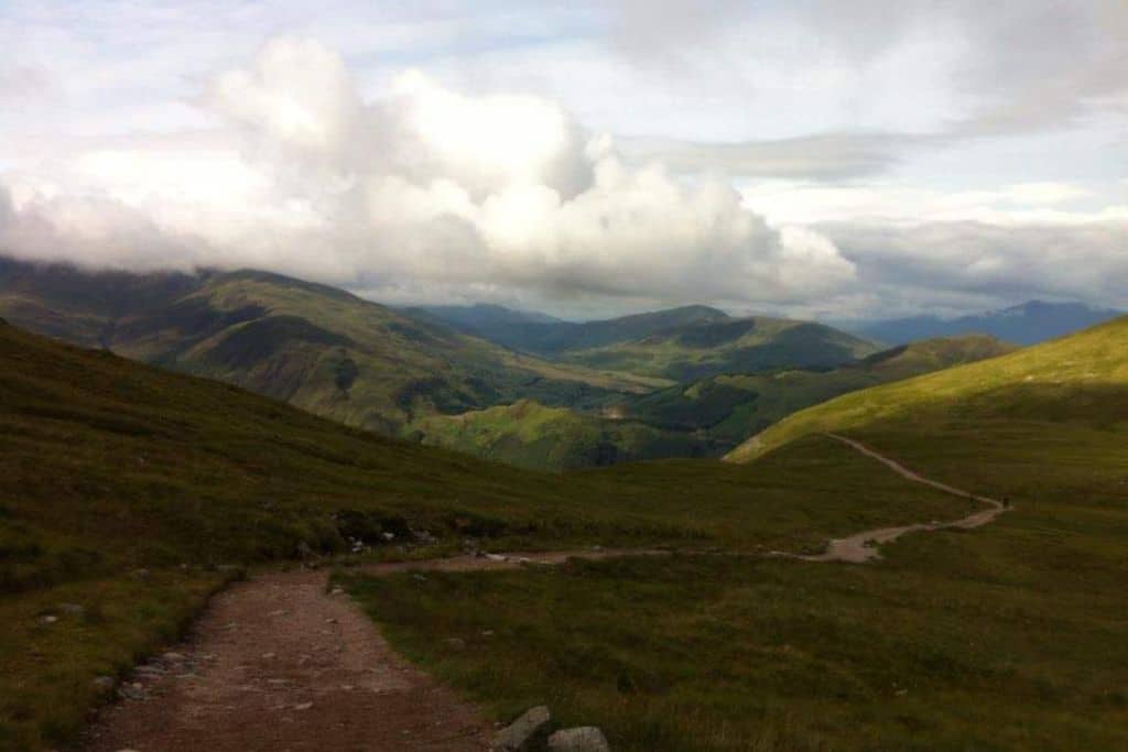 Mountain view down from climbing Ben Nevis of track and rolling green hills with a cloudy sky.