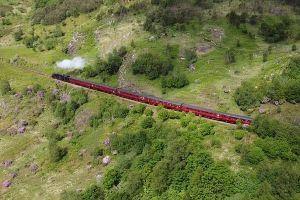 Red steam train puffing steam travelling through green landscape. This is the famous train from Harry Potter.