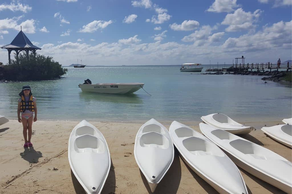 A Kid stood on a beach with 6 white kayaks. Blue sea behind has perfect conditions for kayaking with kids. 