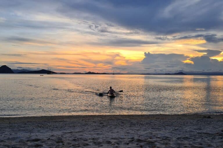 A kid kayaking close to a beach at sunset.