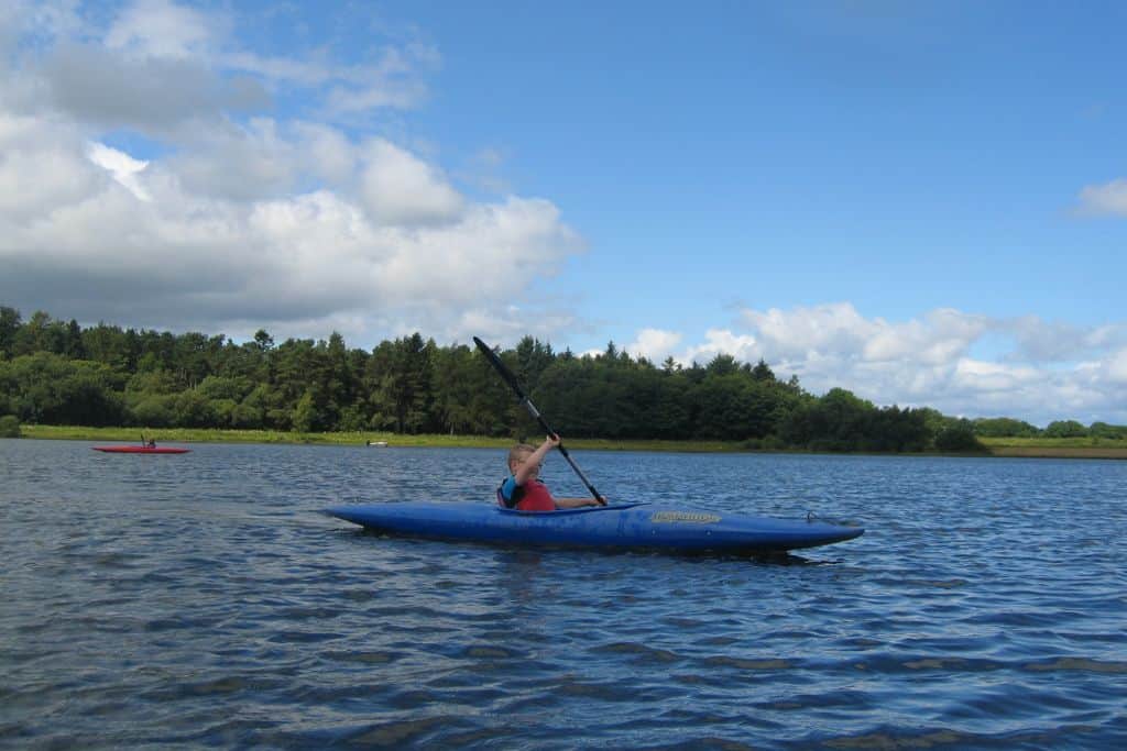 A Kid kayaking on a body of water surrounded by green trees. 