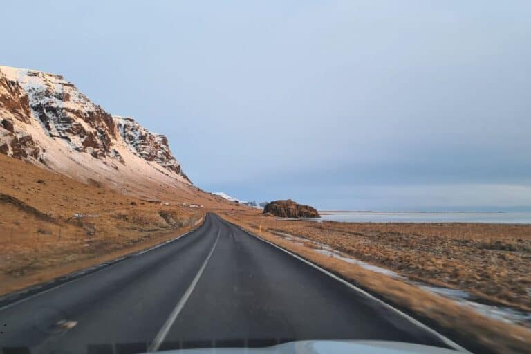 Driving on a tarmac road with snowy hills on one side and sea on the other during an Iceland road trip with kids.