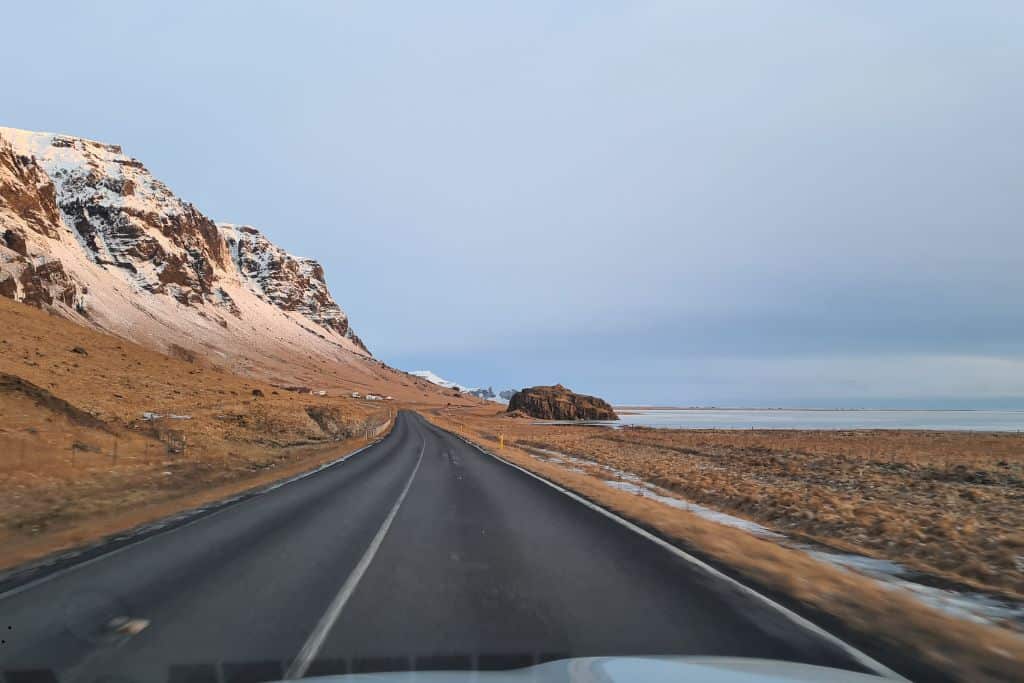 Driving on a tarmac road with snowy hills on one side and sea on the other during an Iceland road trip with kids.