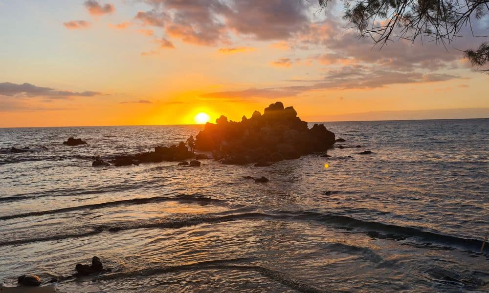 An orange sun setting behind a rock in the sea. A road trip on Big Island offers flexibility to your itinerary.