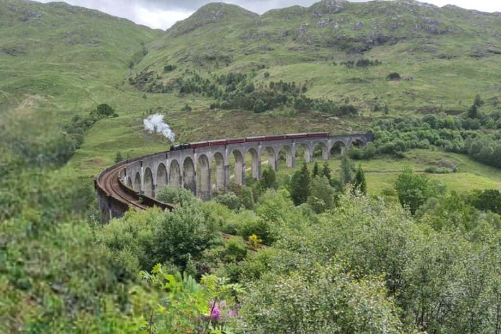 A curved concrete arched viaduct surrounded by green hills. The iconic Jacobite steam train is popular with tourists in Glenfinnan.