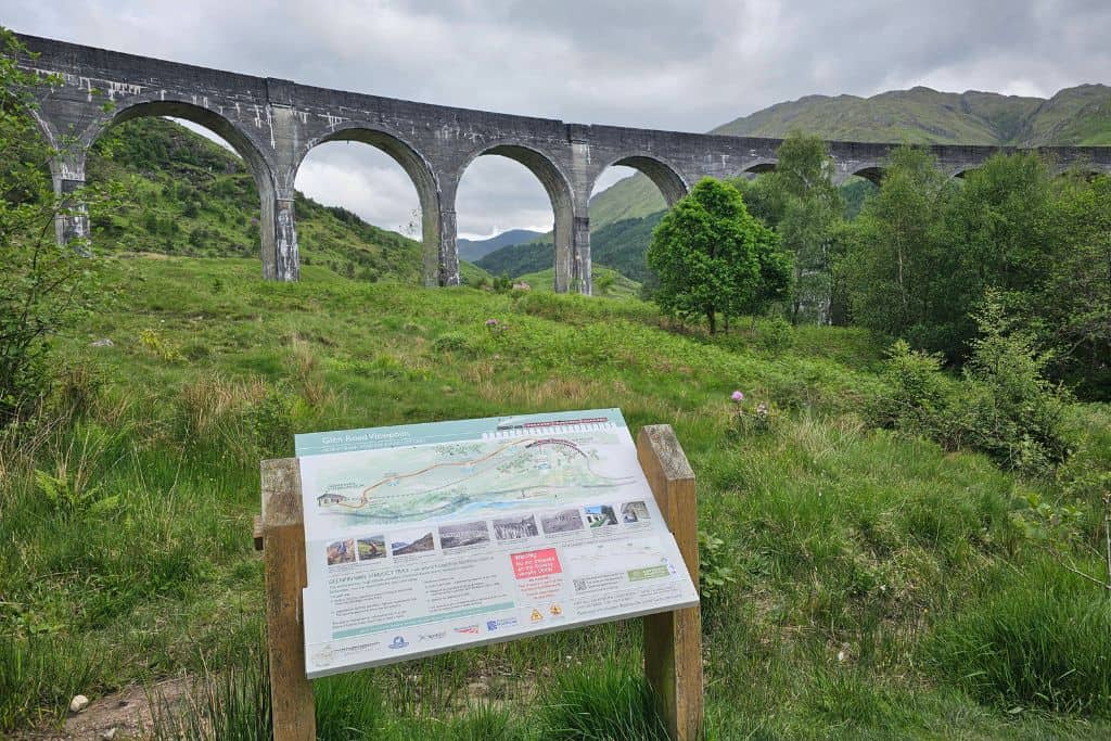 An arched bridge and a viewpoint information sign surrounded by grass. A popular visit in Glenfinnan.