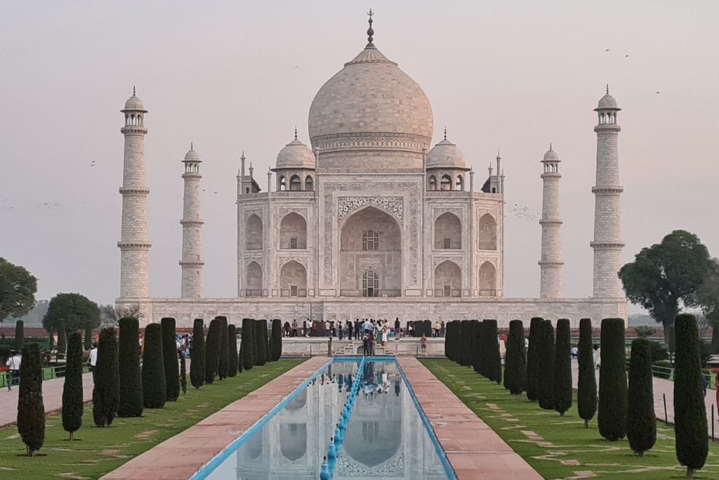 Large white building with manicured garden and pond in front. Visiting the Taj Mahal is on many travellers bucket list.  