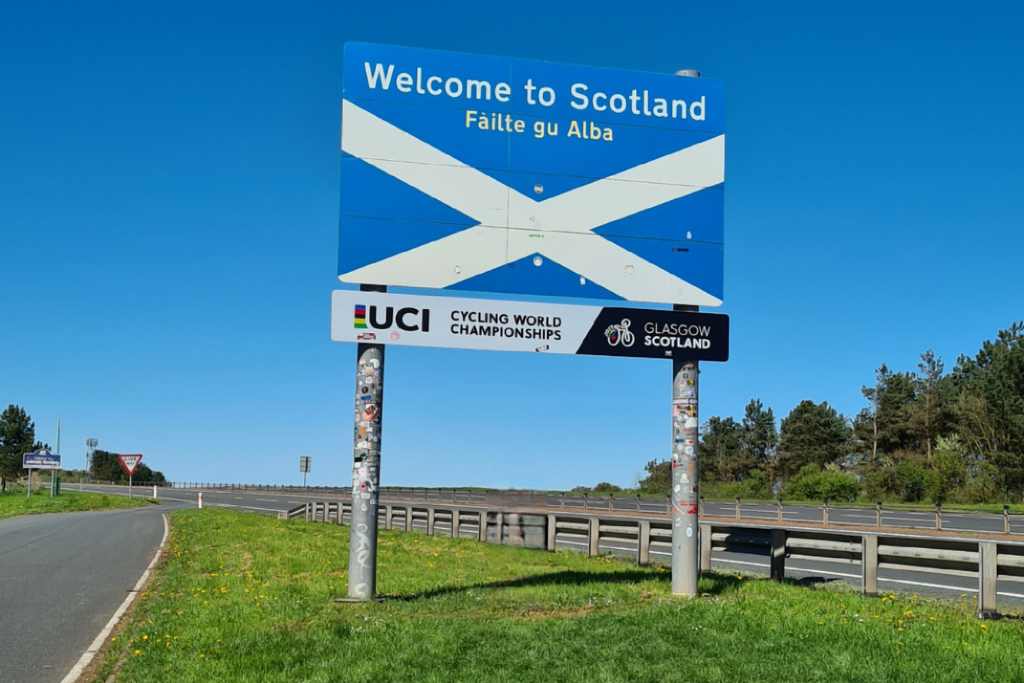 Welcome to Scotland road sign on a blue sign on grass next to a road.