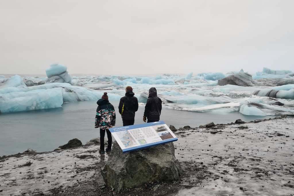 Three kids stood behind a sign looking out into water with iceberg's. A top reason to visit Iceland.