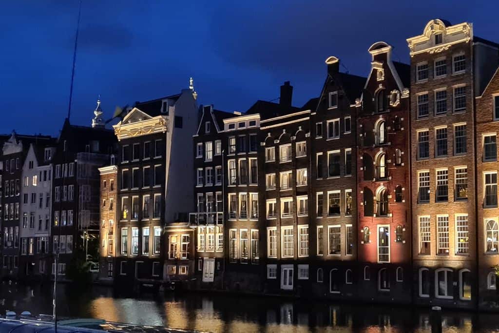 A terrace of narrow colourful town houses lit up at night reflecting into a canal.