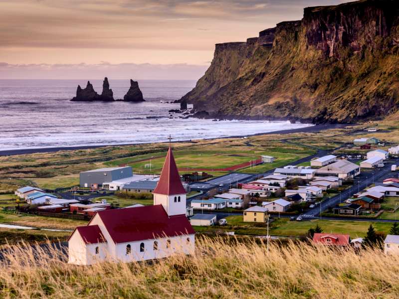 A white and red church overlooking a town with sea stacks in the distance in Vik Iceland.