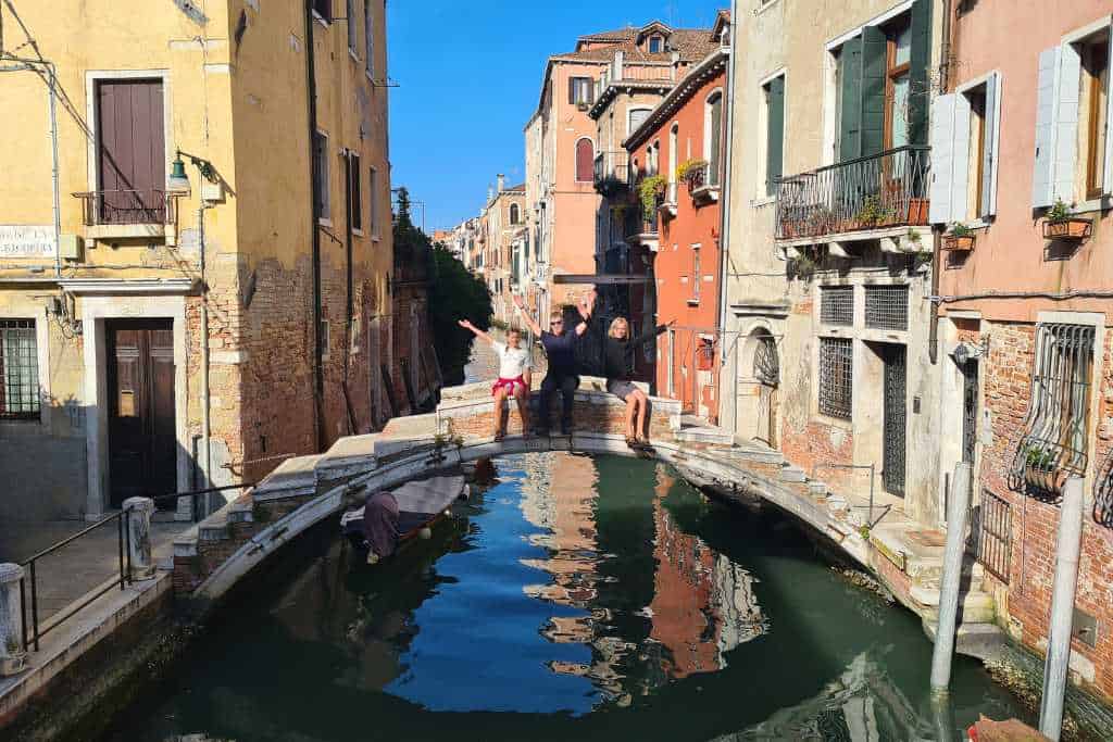 Kids sat on a historic bridge over a canal between buildings in Venice, Italy.