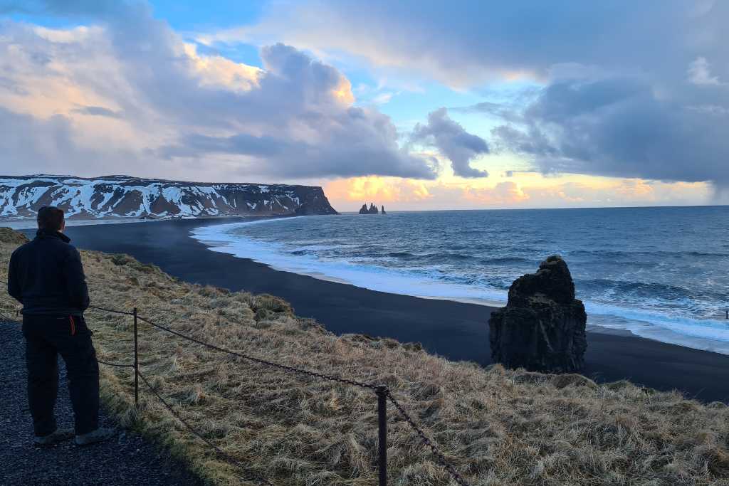 A person looking out over a black sand beach and sea with cliffs and distant pinnacles on a bucket list trip to Iceland.