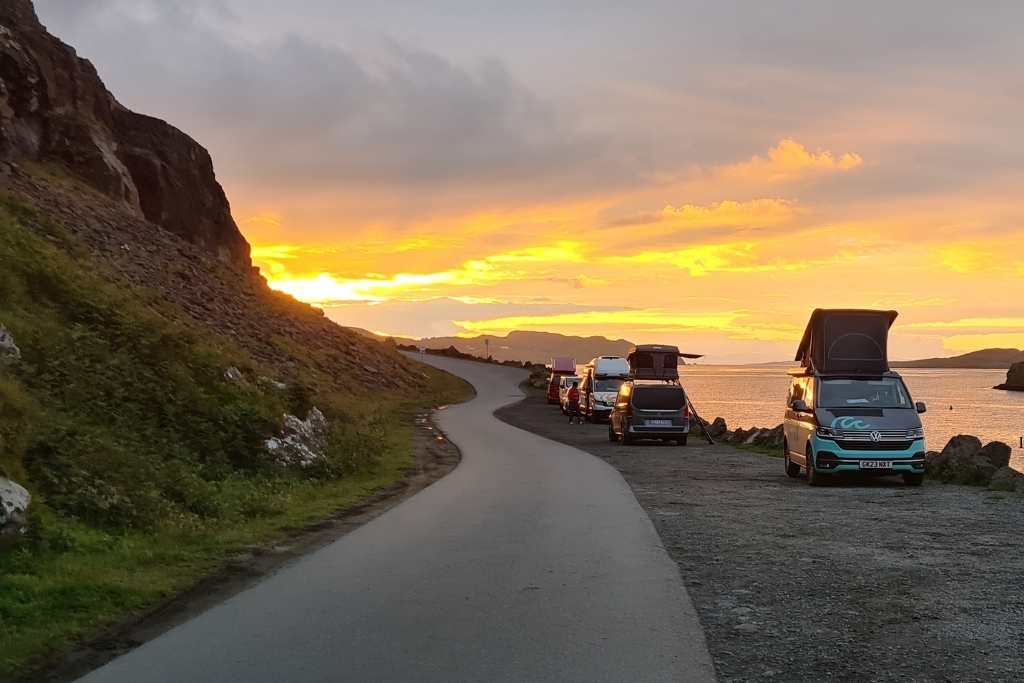 Campers set up at the side of the road with a sunset next to hills and the ocean.