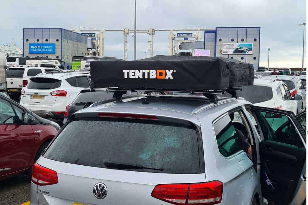 A black TentBox XL fixed to the roof of a silver car at a ferry port.