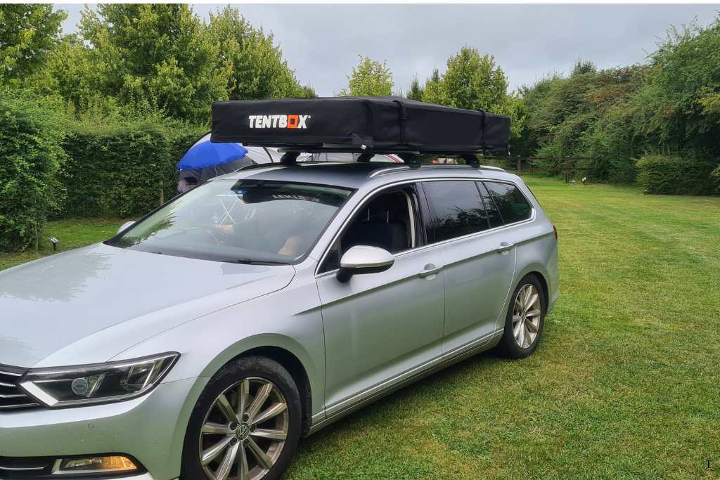 Car with a folded black TentBox on top in a grassy campsite.