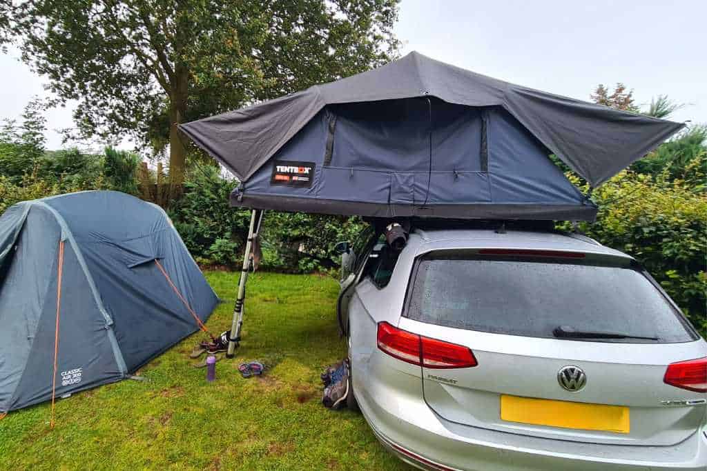 A rooftop tent on a silver car in a campsite next to a tent.