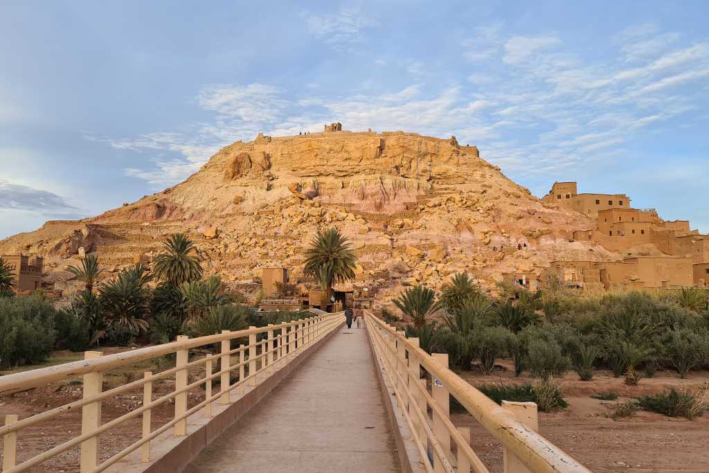 A bridge across a river leading to a City of mud brick houses on a hill. Ben Ait Haddou is a popular traditional Kasbah worth seeing on a Moroccan itinerary.