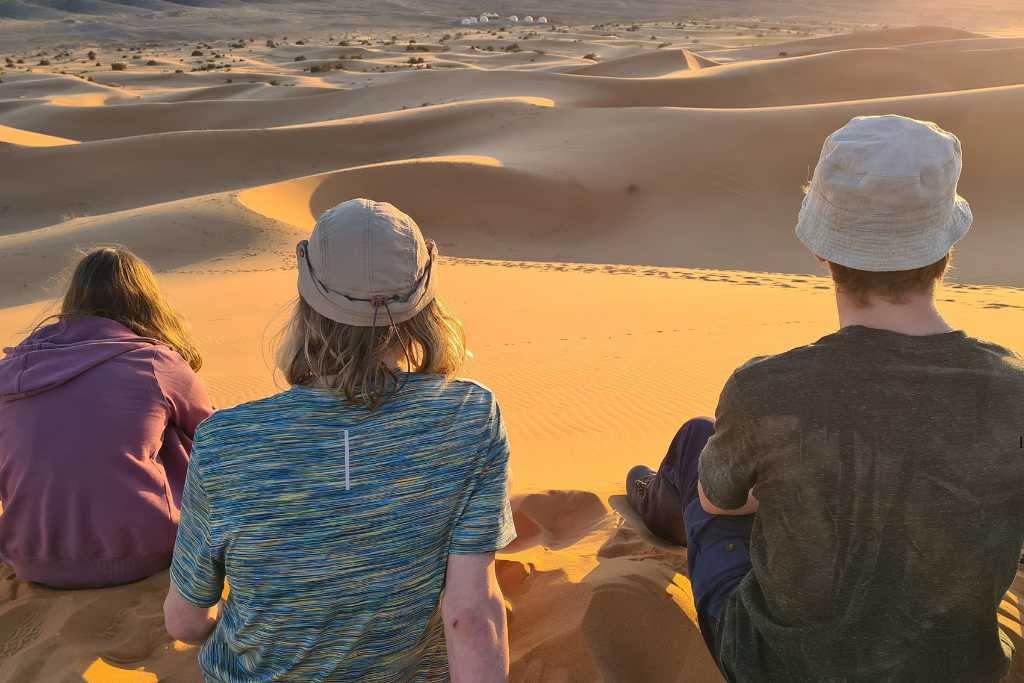 Three kids watching the sun set from a sand dune in the Sahara desert in Morocco.